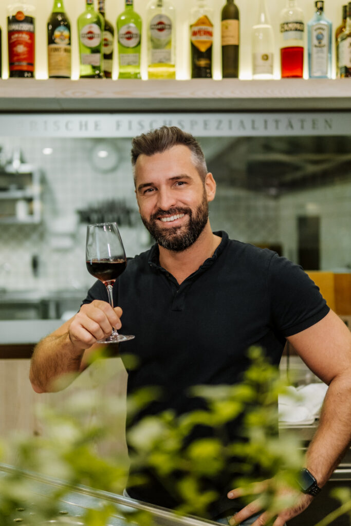 Chefkoch Andreas der Taverna Korfu Marburg hält ein Glas Rotwein und lächelt in die Kamera.